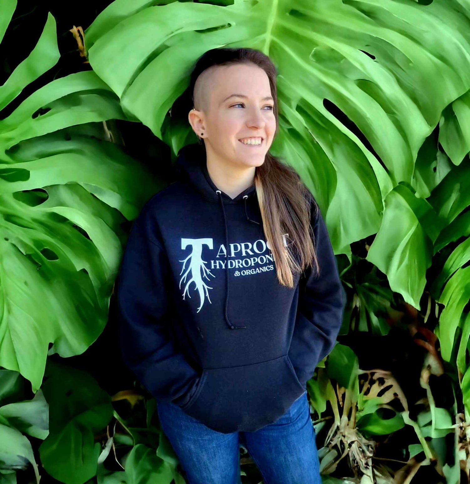 Image: Person in Taproot Hoodie smiling, standing in front of large plant leaf background
